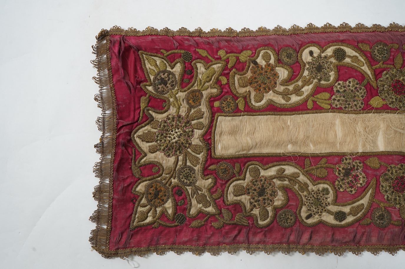 A 19th century Indian red and cream silk runner, heavily embroidered with scrolling motifs, using a variety of stump work and gold wire work stitches, with earlier Mughal influences, 118cm x 46.5cm (not including braid),